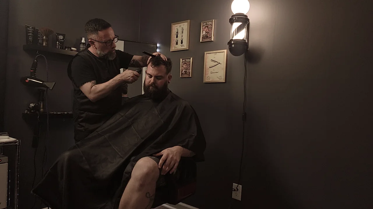 The Barber, wearing glasses and a black shirt, carefully shaping a client's haircut